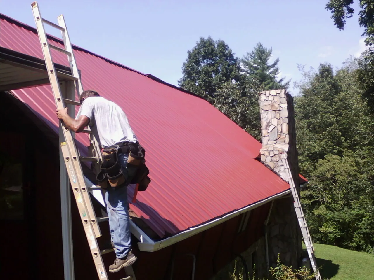 Roofer on a ladder beside a red metal roof and stone chimney — Soffit & Fascia Repair by Roofing Brothers in Pitcairn